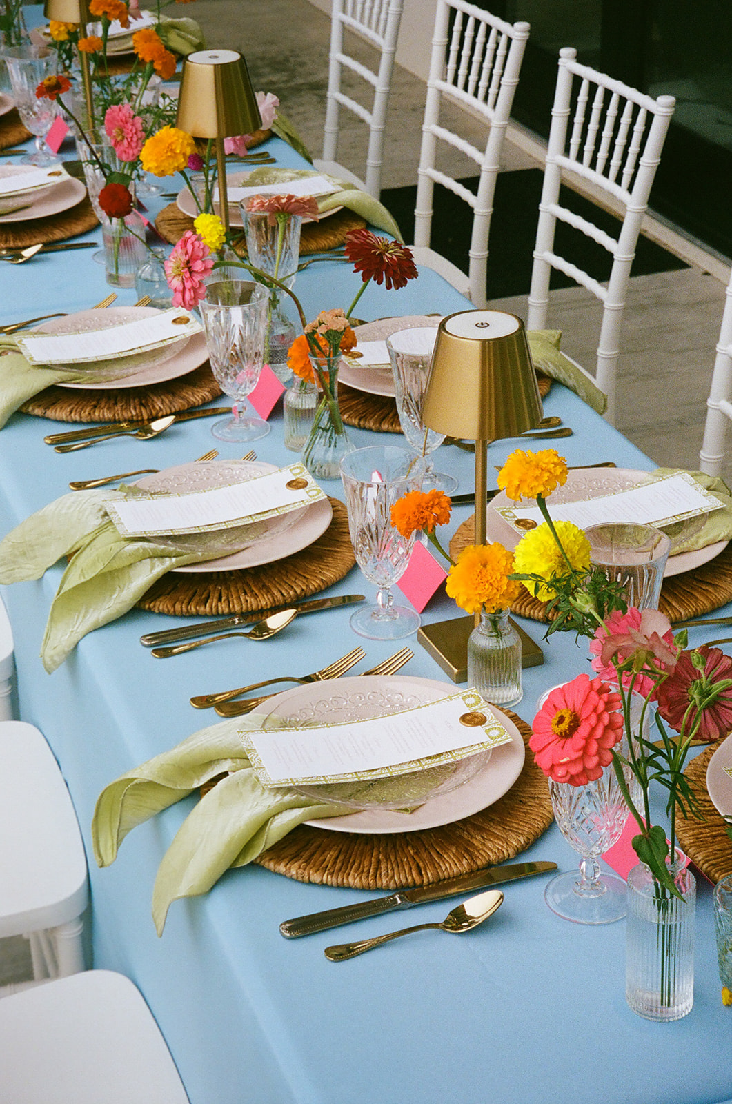 A long dining table with a light blue tablecloth, gold cups, glassware, and colorful flower arrangements is set outdoors beside a stone wall. | Unique wedding design