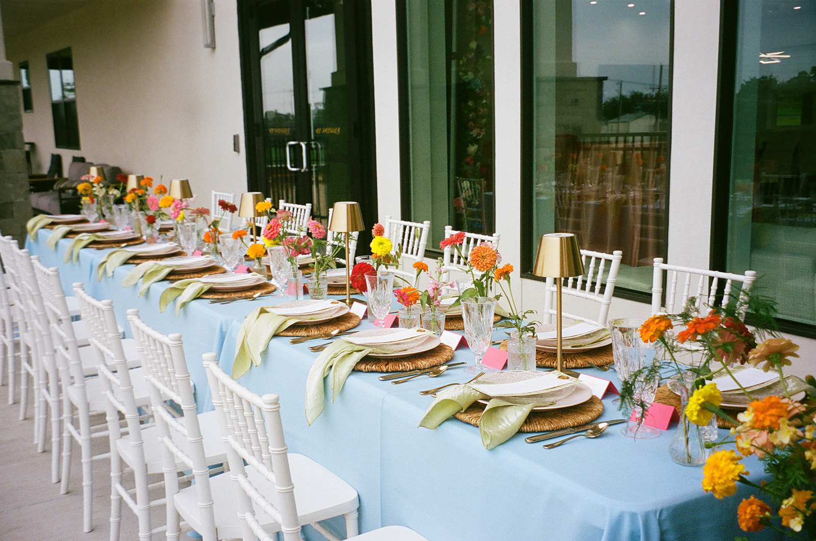 A long dining table with a light blue tablecloth, gold cups, glassware, and colorful flower arrangements is set outdoors beside a stone wall. | Unique wedding design
