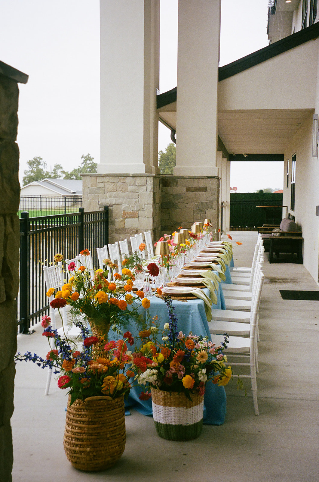 A long dining table with a light blue tablecloth, gold cups, glassware, and colorful flower arrangements is set outdoors beside a stone wall. | Unique wedding design