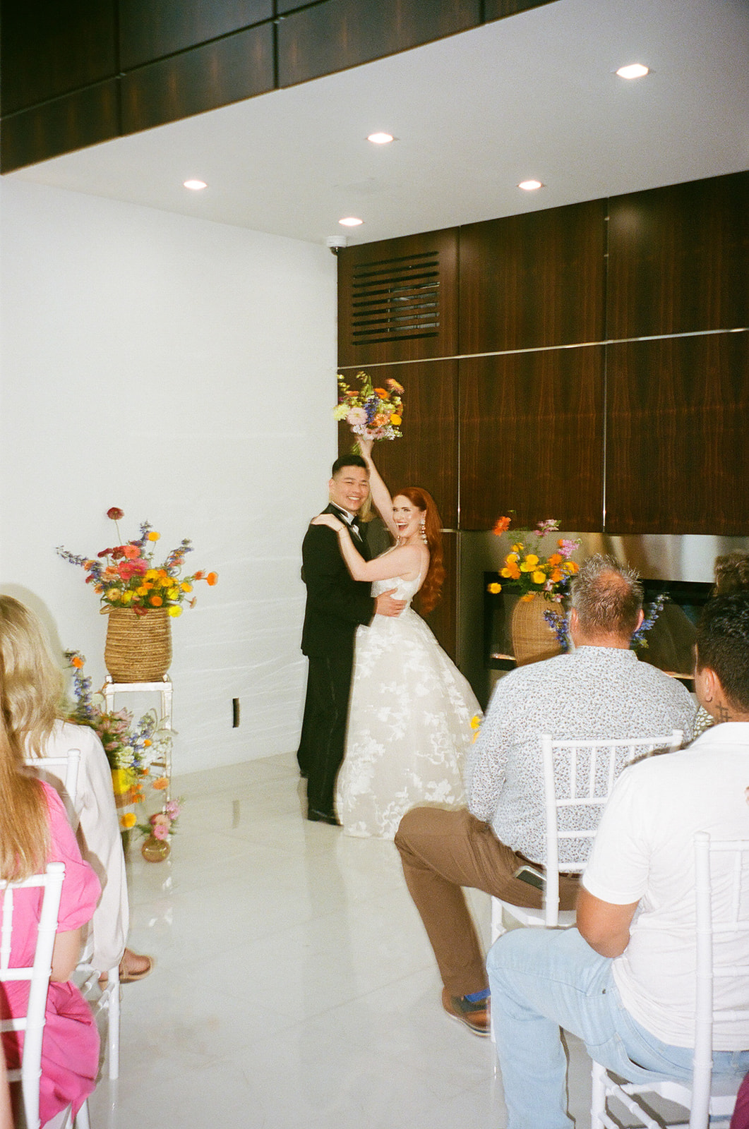 A couple stands with an officiant exchanging vows in a modern indoor wedding ceremony, with guests seated and floral arrangements on display.