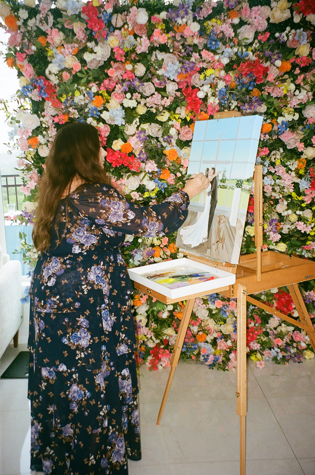 A woman with long brown hair paints on a canvas set on an easel, standing in front of a wall covered with colorful flowers.