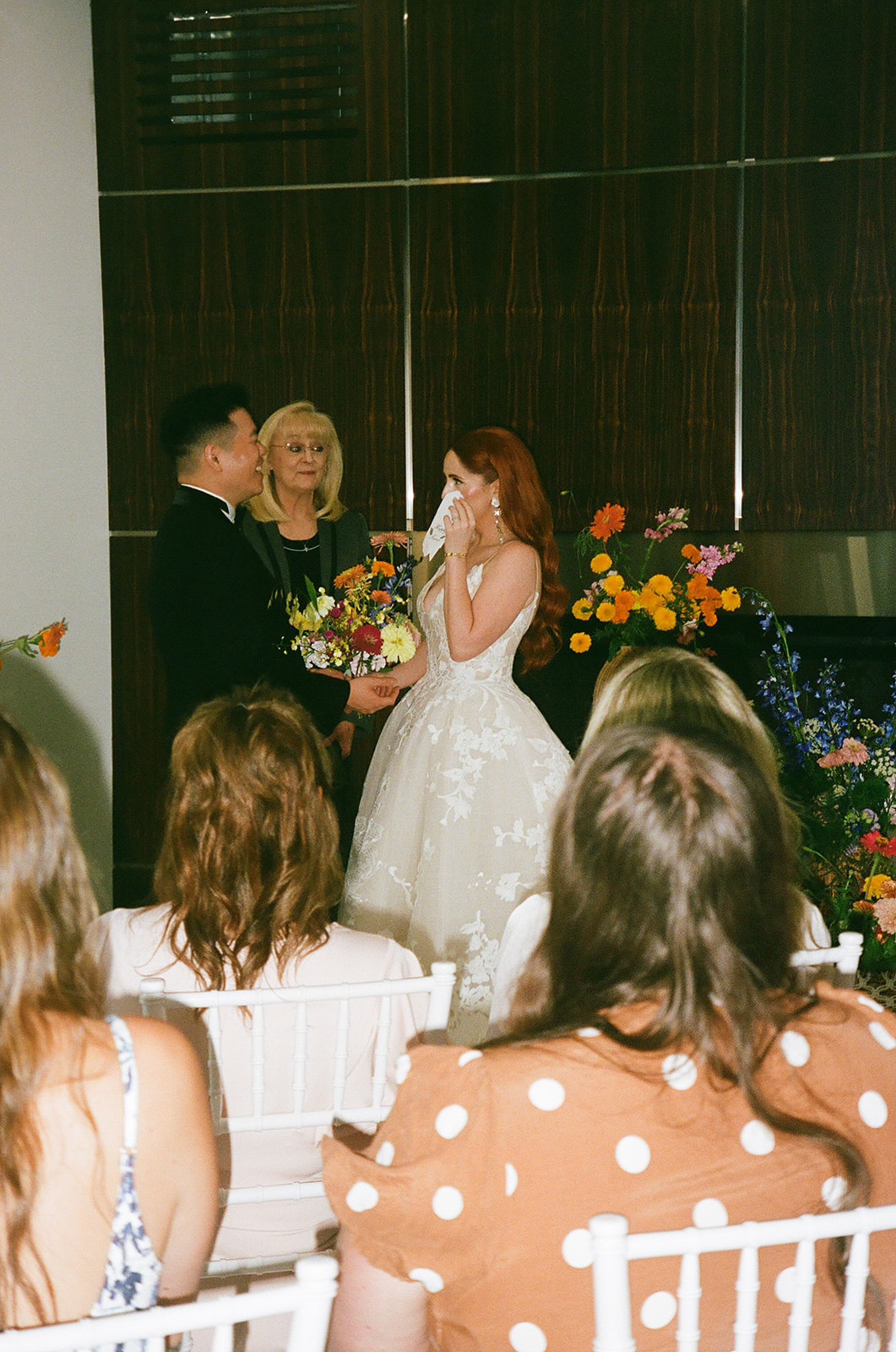 A couple stands with an officiant exchanging vows in a modern indoor wedding ceremony, with guests seated and floral arrangements on display.