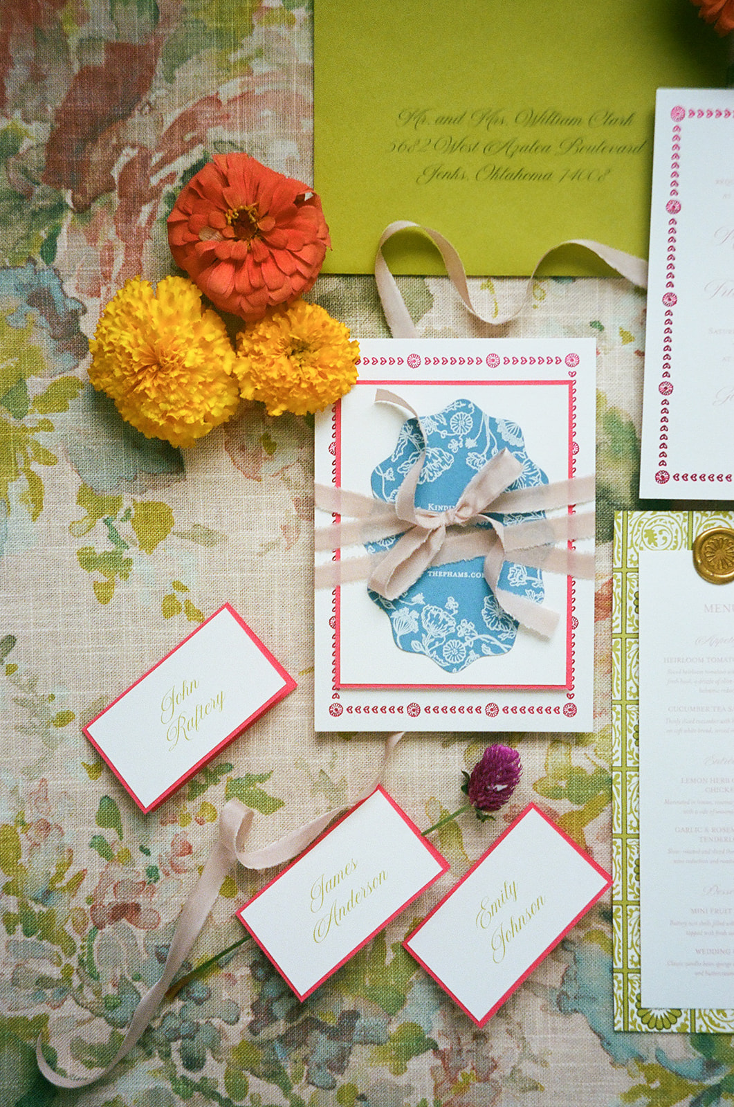 Flat lay of floral-themed stationery, place cards, a ribbon-tied invitation with a blue wax seal, yellow and orange flowers, and a green envelope on a floral fabric background. | unique wedding design