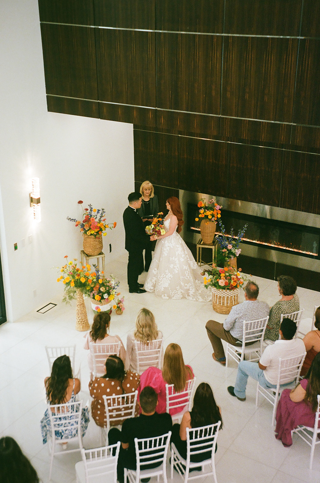A couple stands with an officiant exchanging vows in a modern indoor wedding ceremony, with guests seated and floral arrangements on display.
