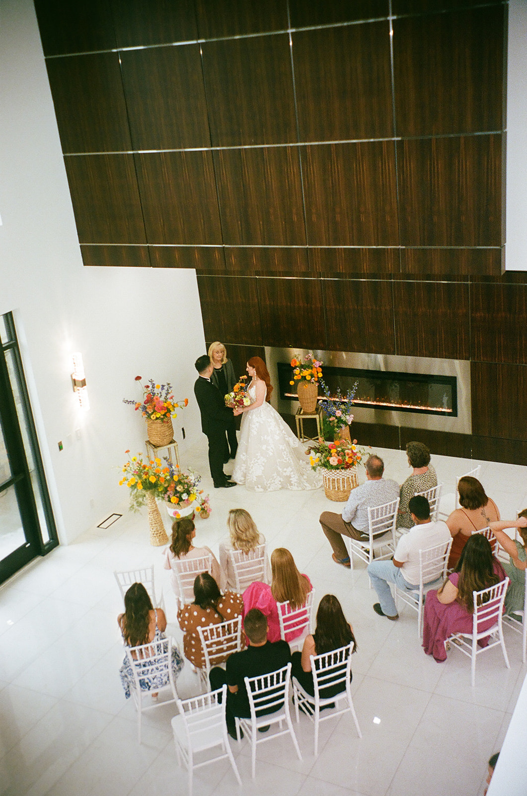 A couple stands with an officiant exchanging vows in a modern indoor wedding ceremony, with guests seated and floral arrangements on display.