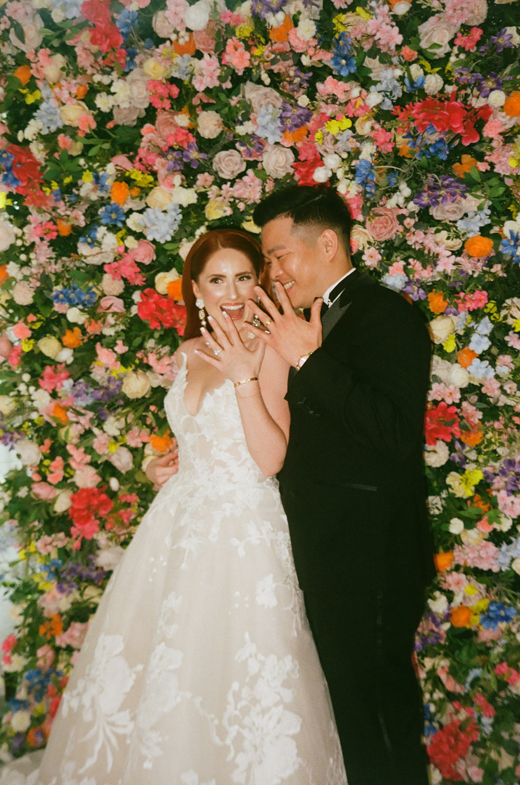A bride and groom stand in front of a colorful flower wall, smiling and showing their wedding rings to the camera.