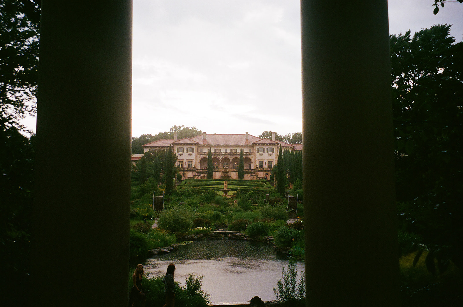 A couple dressed in white holds hands on a stone terrace in front of a large building with tall windows and potted plants.