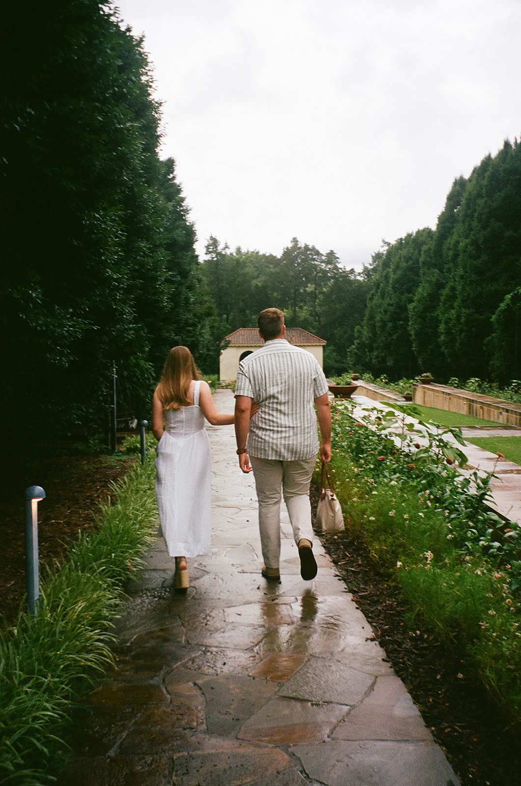 Two people dressed in white walk up stone steps toward a small white pavilion with columns, surrounded by greenery.