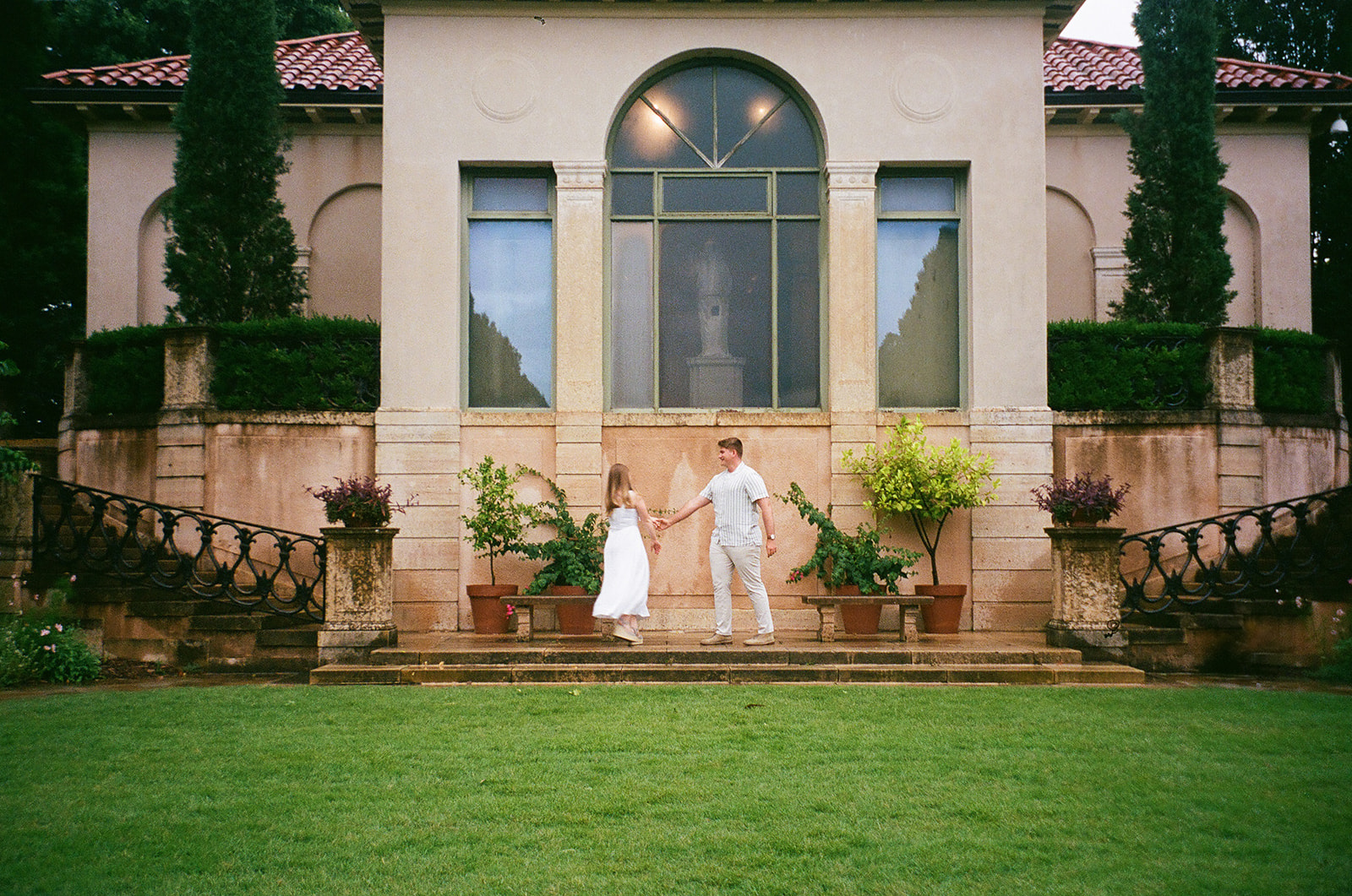 A couple dressed in white holds hands on a stone terrace in front of a large building with tall windows and potted plants.