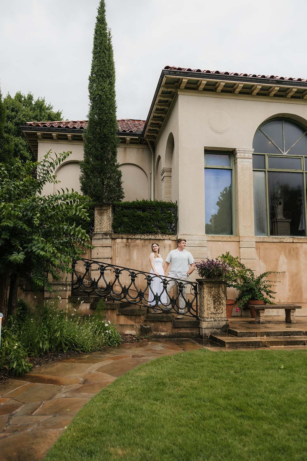 A bride and groom stand together on the steps of a large, elegant villa with tall windows and landscaped greenery at the Philbrook Museum in Tulsa Oklahoma