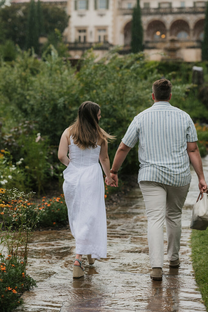 A couple walks hand in hand down a wet garden path, surrounded by greenery and flowers, with a large building in the background.