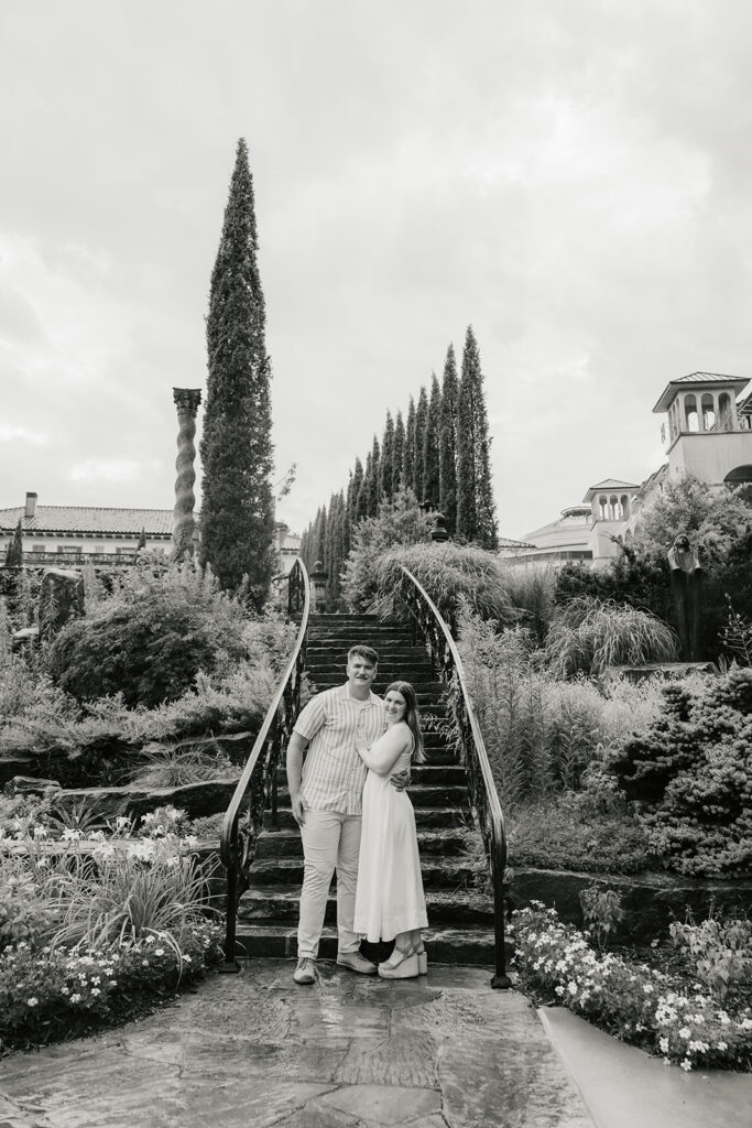 A couple stands together on a stone staircase in a landscaped garden, with tall trees and buildings in the background.