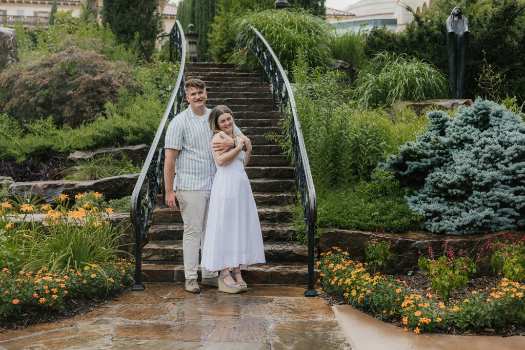 A couple stands together on a stone staircase in a landscaped garden, with tall trees and buildings in the background.