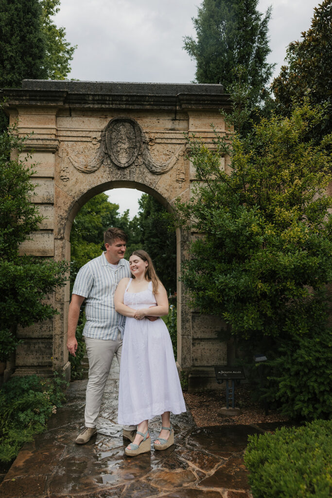 A man and woman stand together in front of a stone archway in a garden, with greenery surrounding them.