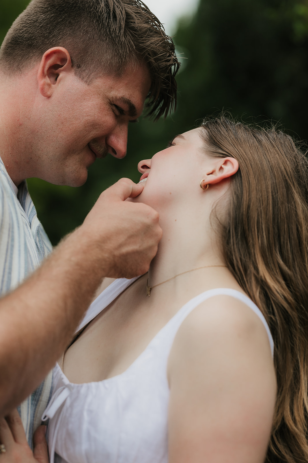 A woman in a white dress and a man in a striped shirt kiss beside stone steps in a formal garden on a cloudy day.
