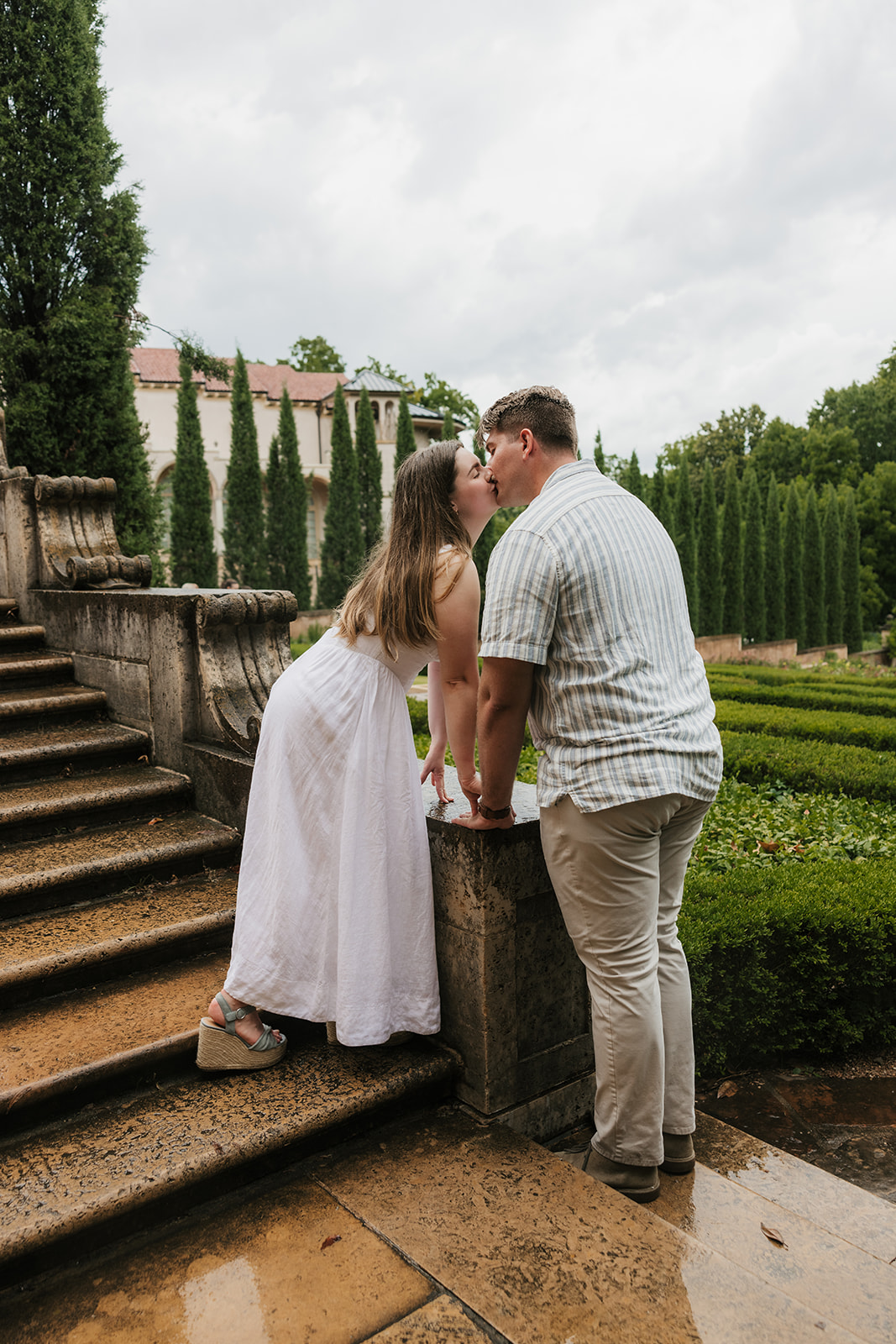A woman in a white dress and a man in a striped shirt kiss beside stone steps in a formal garden on a cloudy day.