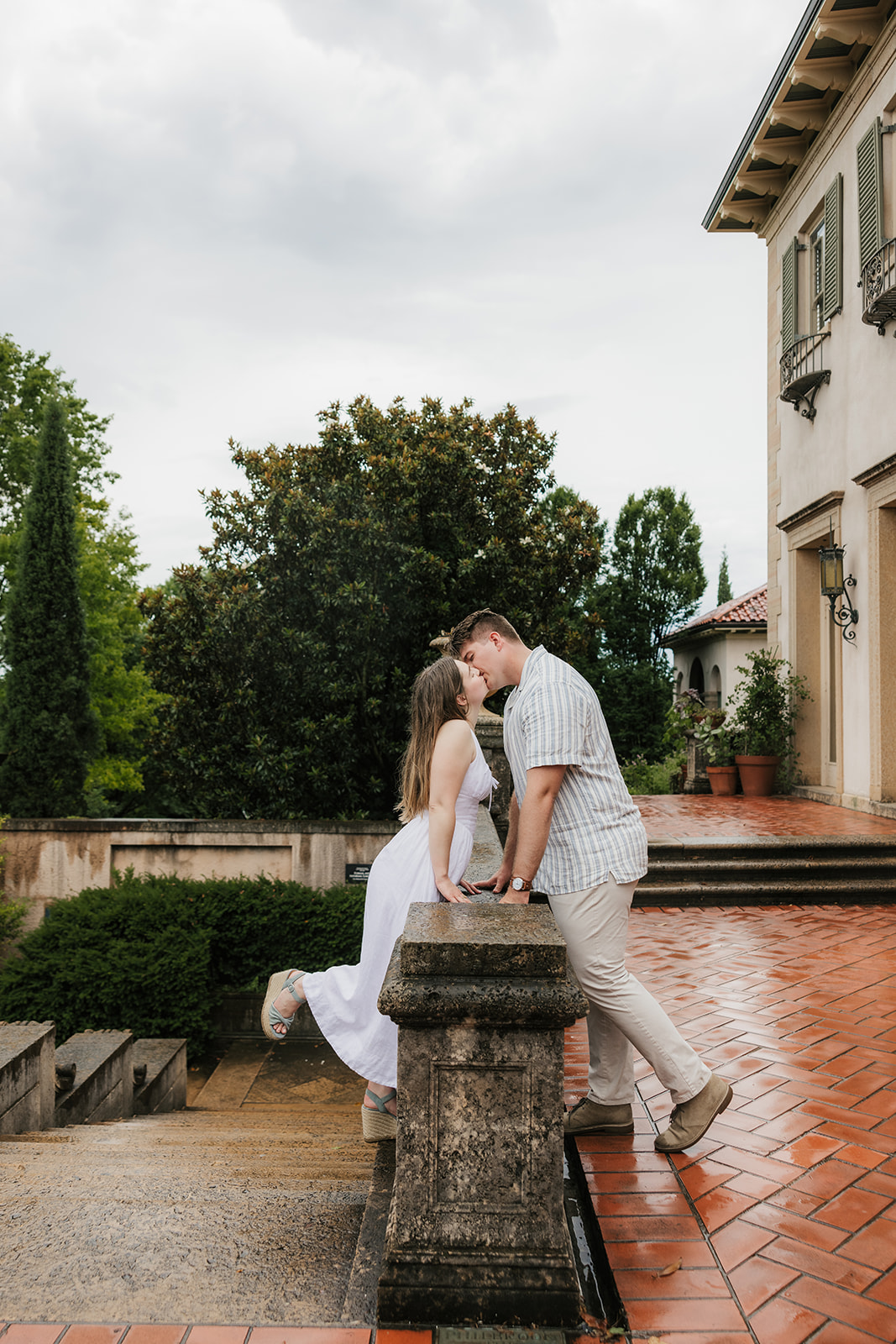 A woman in a white dress and a man in a striped shirt kiss beside stone steps in a formal garden on a cloudy day.