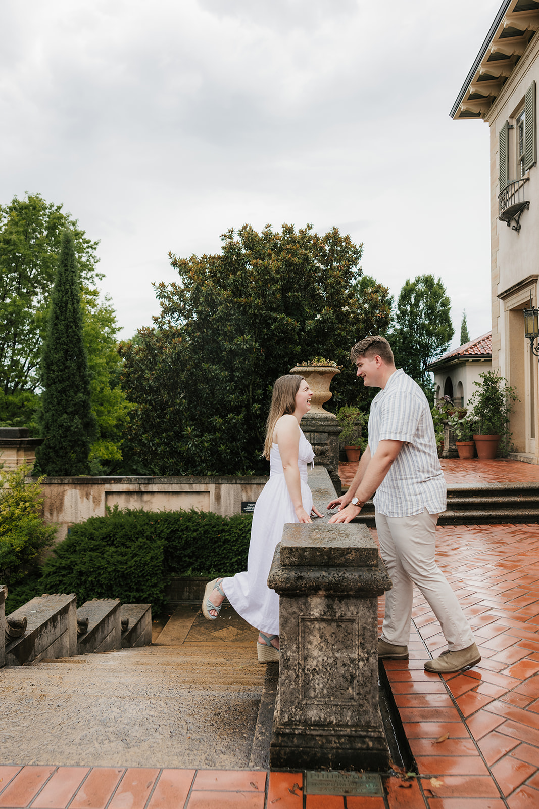A woman in a white dress and a man in a striped shirt kiss beside stone steps in a formal garden on a cloudy day.
