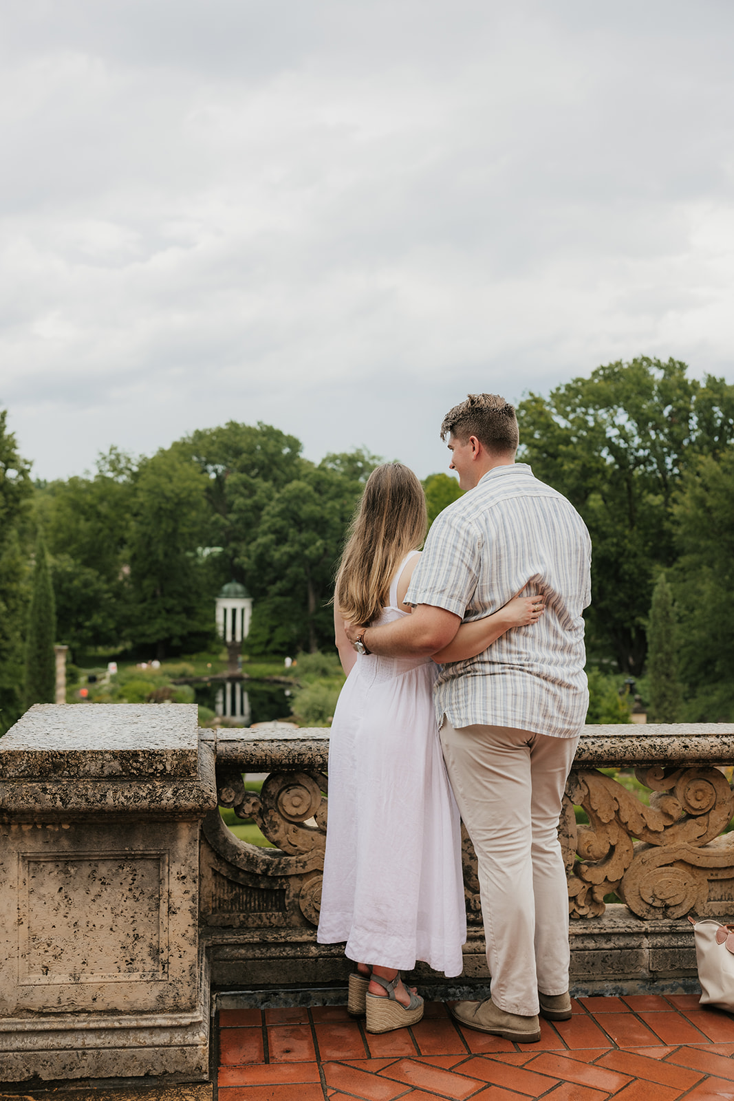 A woman in a white dress and a man in a striped shirt kiss beside stone steps in a formal garden on a cloudy day.
