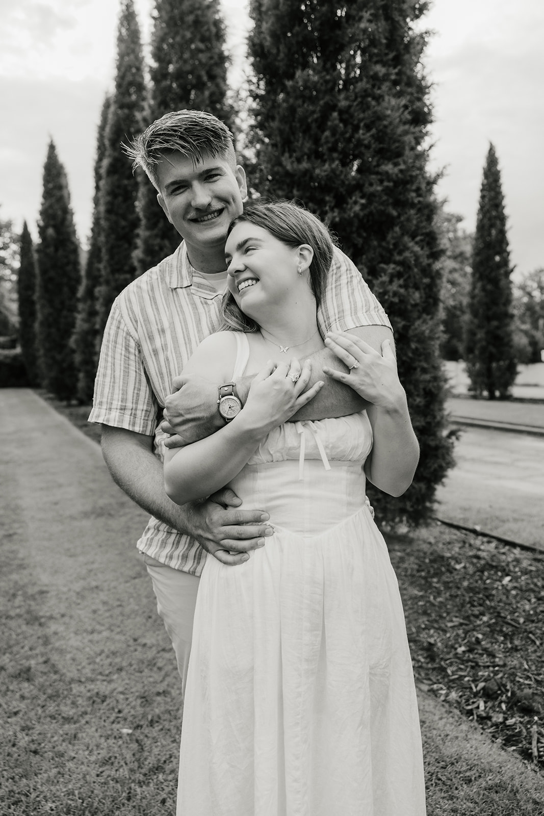 A couple stands outdoors, smiling and embracing each other. Tall trees line the background in a landscaped garden setting. The image is in black and white.