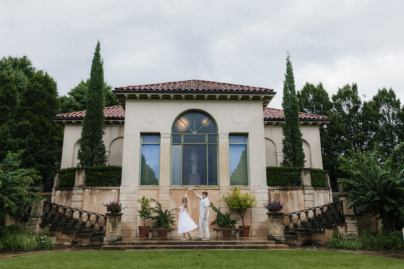 A bride and groom stand together on the steps of a large, elegant villa with tall windows and landscaped greenery at the Philbrook Museum in Tulsa Oklahoma