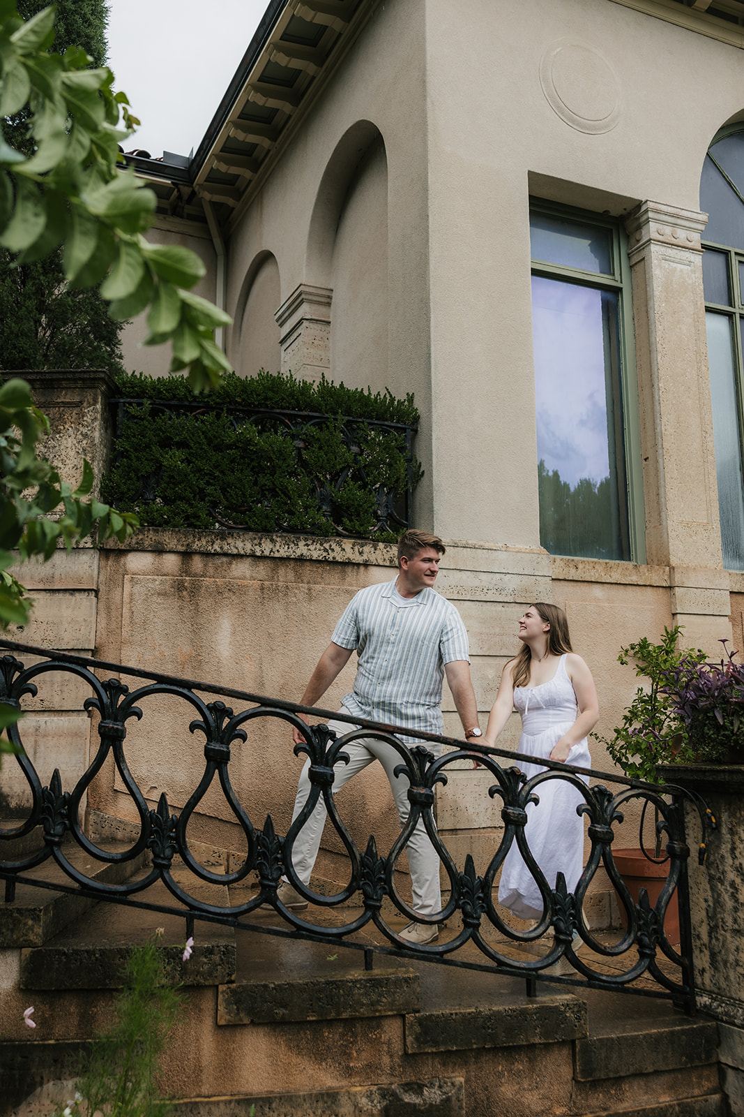 A bride and groom stand together on the steps of a large, elegant villa with tall windows and landscaped greenery at the Philbrook Museum in Tulsa Oklahoma