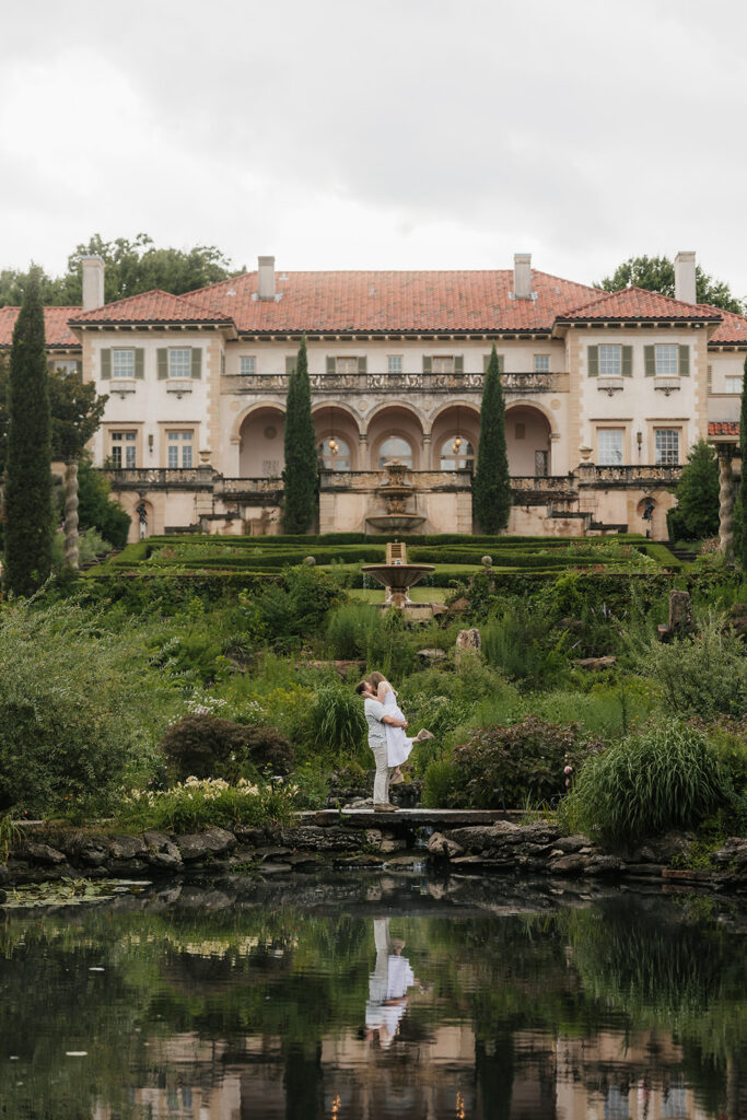 A couple stands by a pond in front of a large, ornate mansion with terraced gardens and tall cypress trees.