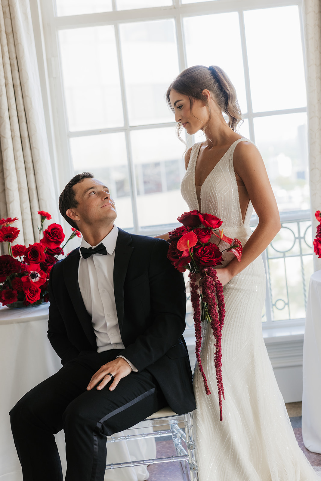 A woman in a white wedding dress holding a bouquet of red flowers stands beside a seated man in a black tuxedo, with red floral arrangements on tables nearby.