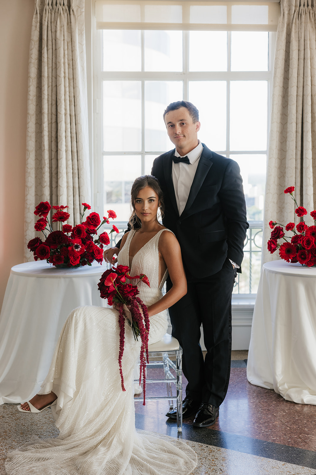 A woman in a white wedding dress holding a bouquet of red flowers stands beside a seated man in a black tuxedo, with red floral arrangements on tables nearby.