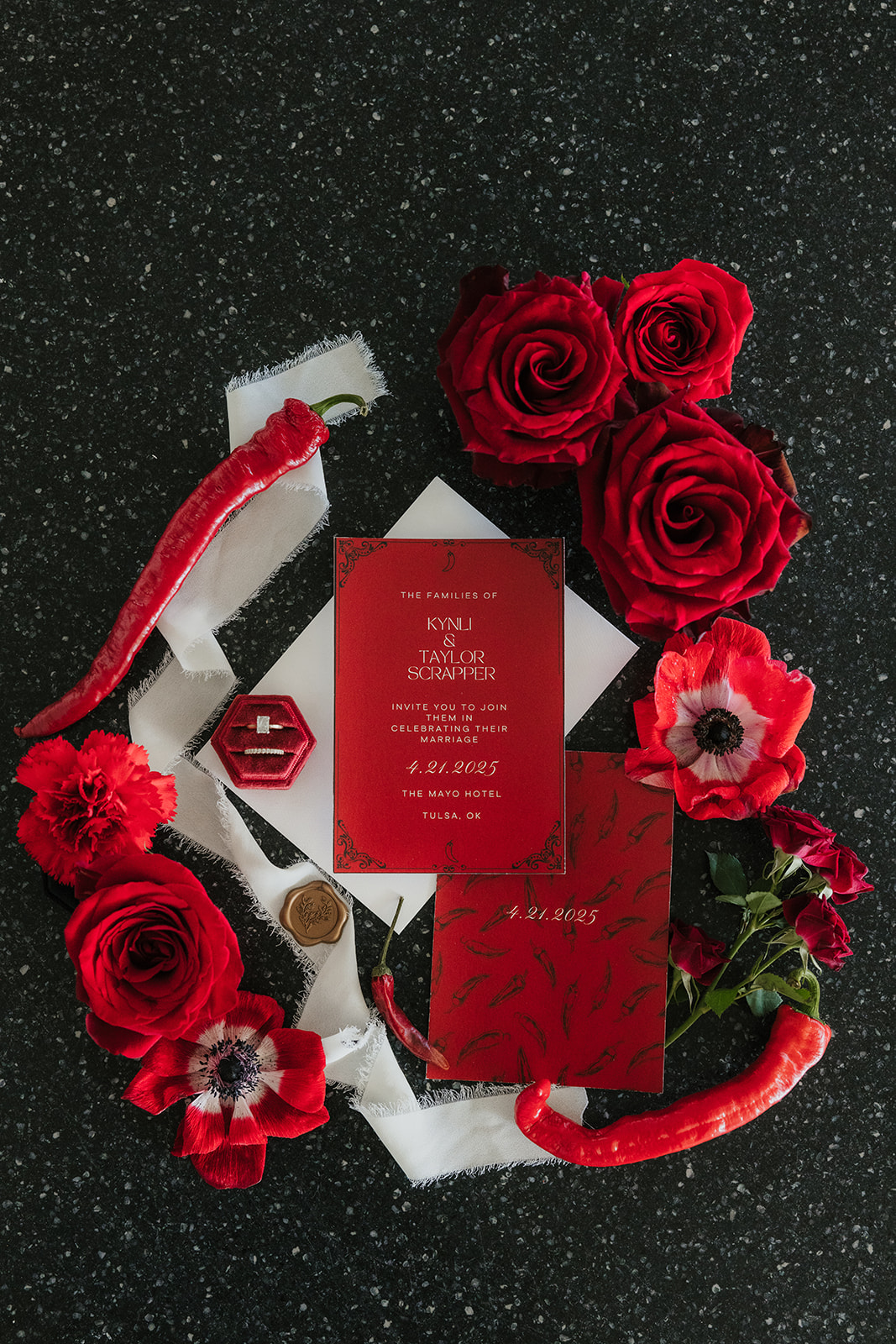 Red wedding invitation suite with red flowers, red chili peppers, a ring box, and white ribbon arranged on a dark speckled surface.