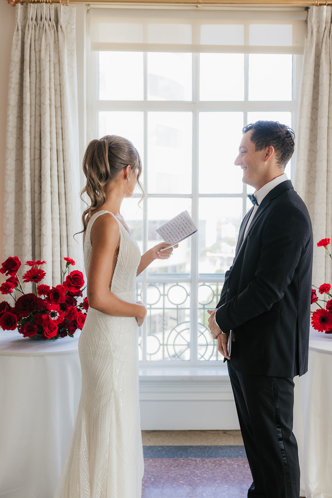 A bride in a white dress holds a piece of paper and smiles at a groom in a dark suit during a wedding ceremony indoors for a wedding at the mayo hotel
