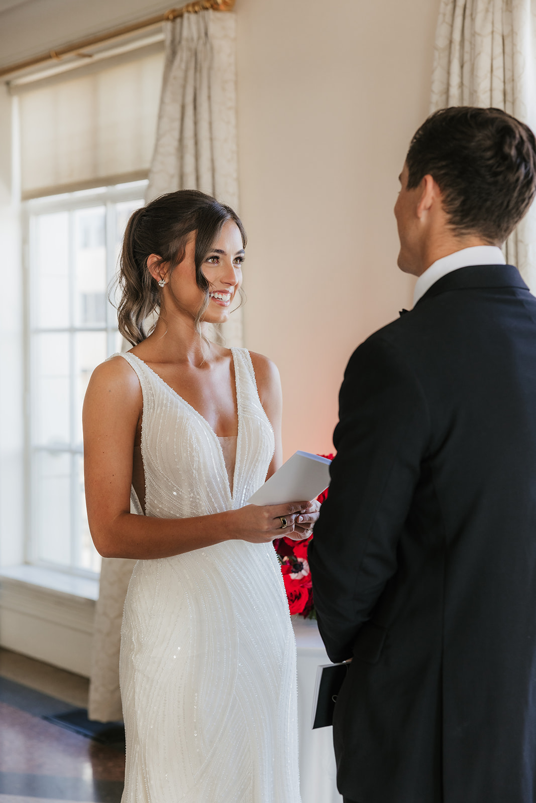 A bride in a white dress holds a piece of paper and smiles at a groom in a dark suit during a wedding ceremony indoors for a wedding at the mayo hotel