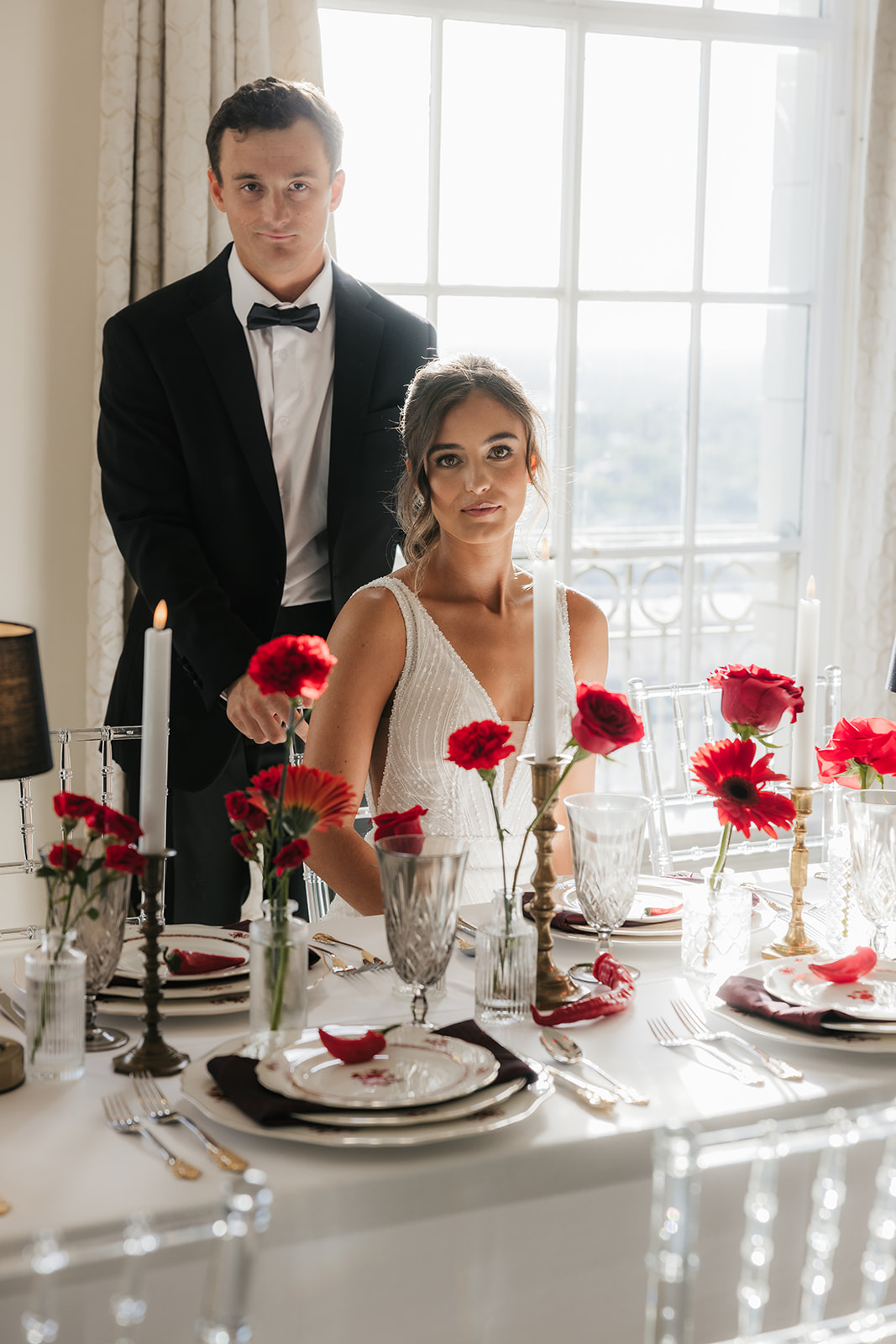 A woman in a white wedding dress holding a bouquet of red flowers stands beside a seated man in a black tuxedo, with red floral arrangements on tables nearby for a wedding at the mayo hotel