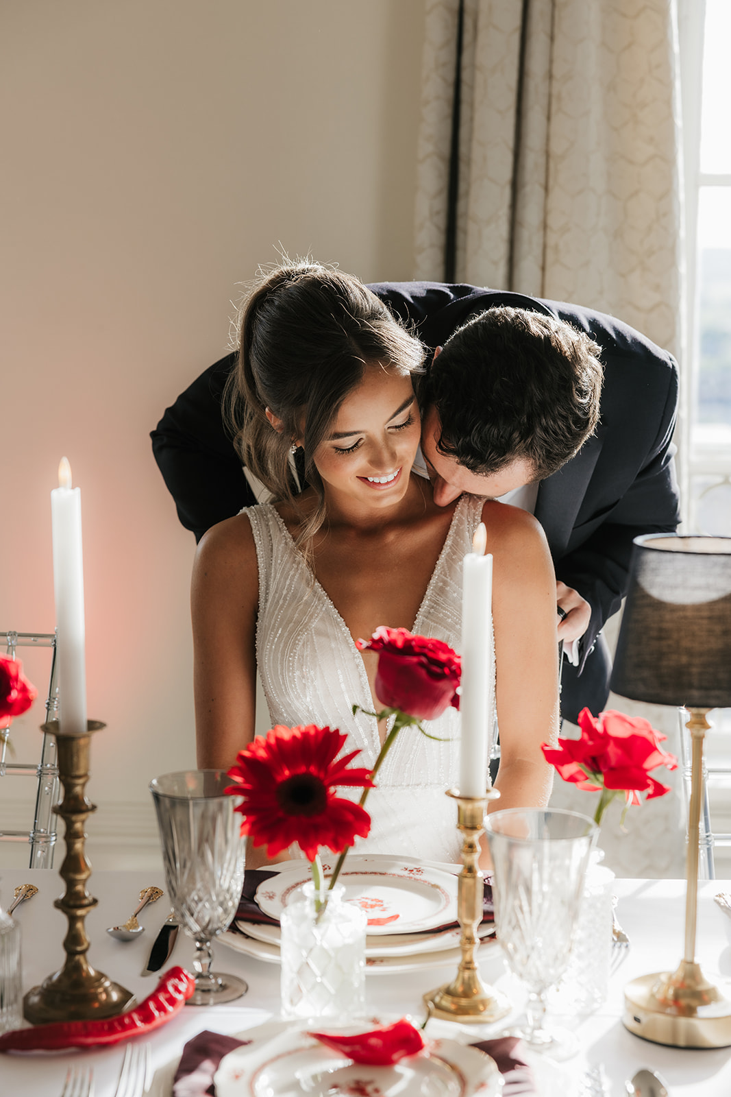 A woman in a white wedding dress holding a bouquet of red flowers stands beside a seated man in a black tuxedo, with red floral arrangements on tables nearby for a wedding at the mayo hotel