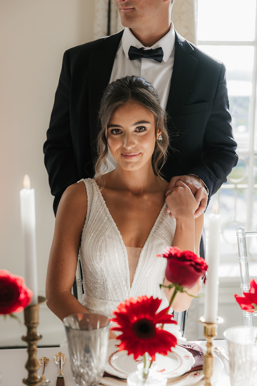 A woman in a white wedding dress holding a bouquet of red flowers stands beside a seated man in a black tuxedo, with red floral arrangements on tables nearby.
