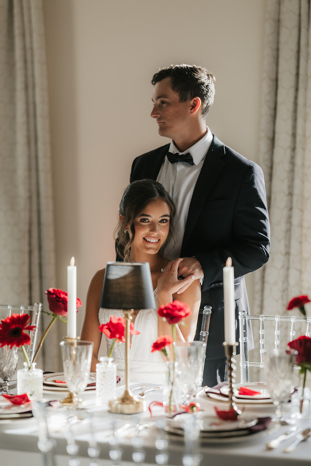 A woman in a white wedding dress holding a bouquet of red flowers stands beside a seated man in a black tuxedo, with red floral arrangements on tables nearby.