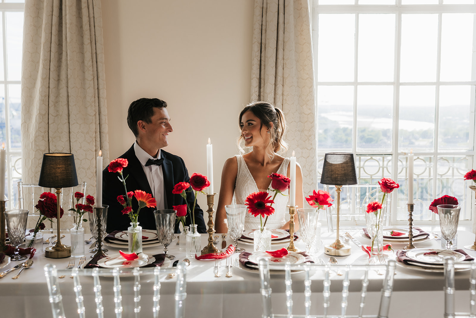 A woman in a white wedding dress holding a bouquet of red flowers stands beside a seated man in a black tuxedo, with red floral arrangements on tables nearby for a wedding at the mayo hotel