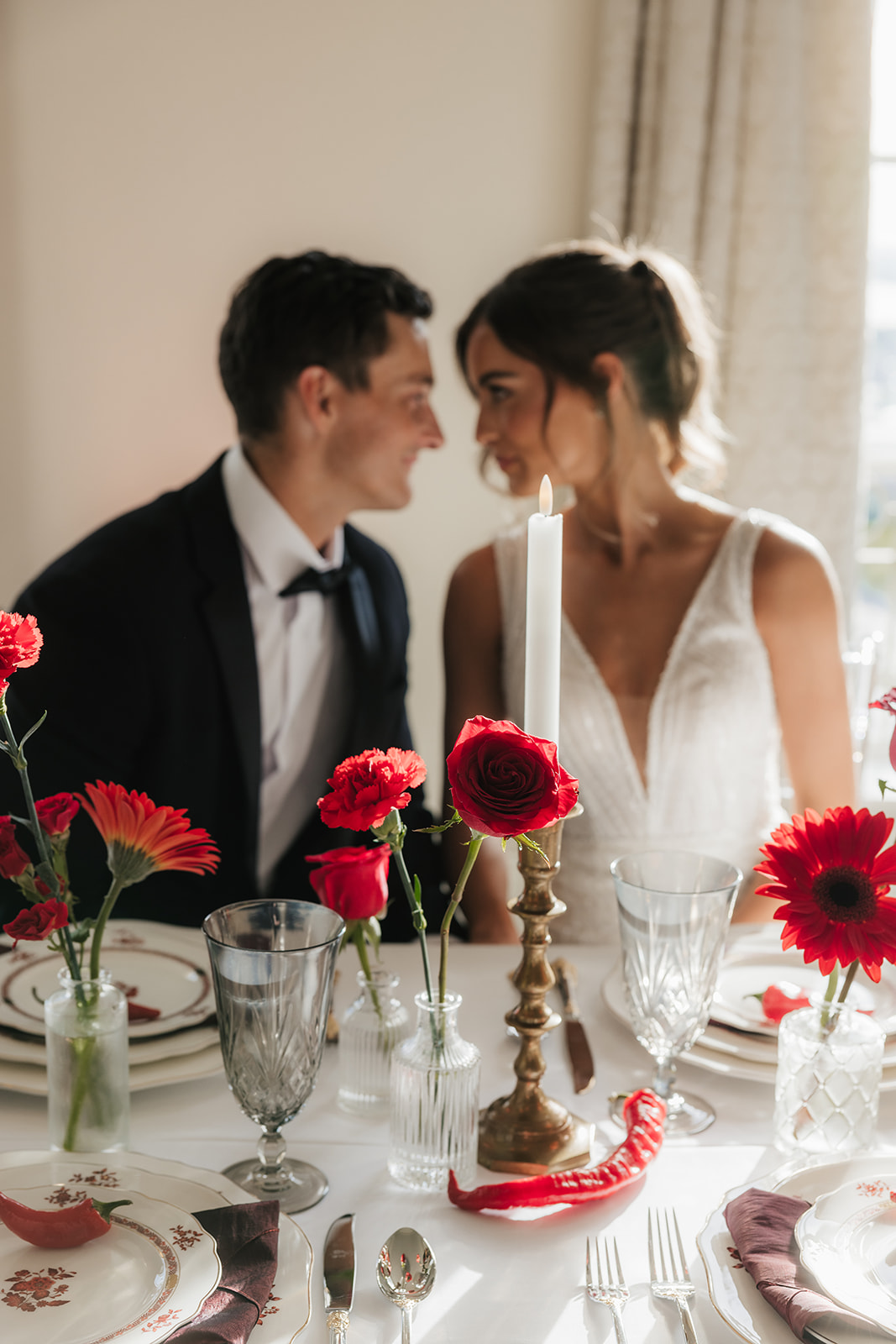 A woman in a white wedding dress holding a bouquet of red flowers stands beside a seated man in a black tuxedo, with red floral arrangements on tables nearby for a wedding at the mayo hotel