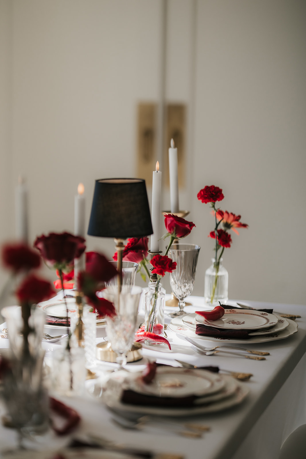 A formal table setting with stacked plates, crystal glassware, gold utensils, red napkin, a red chili pepper on the top plate, and red roses in vases on a white tablecloth for a Mayo Hotel wedding