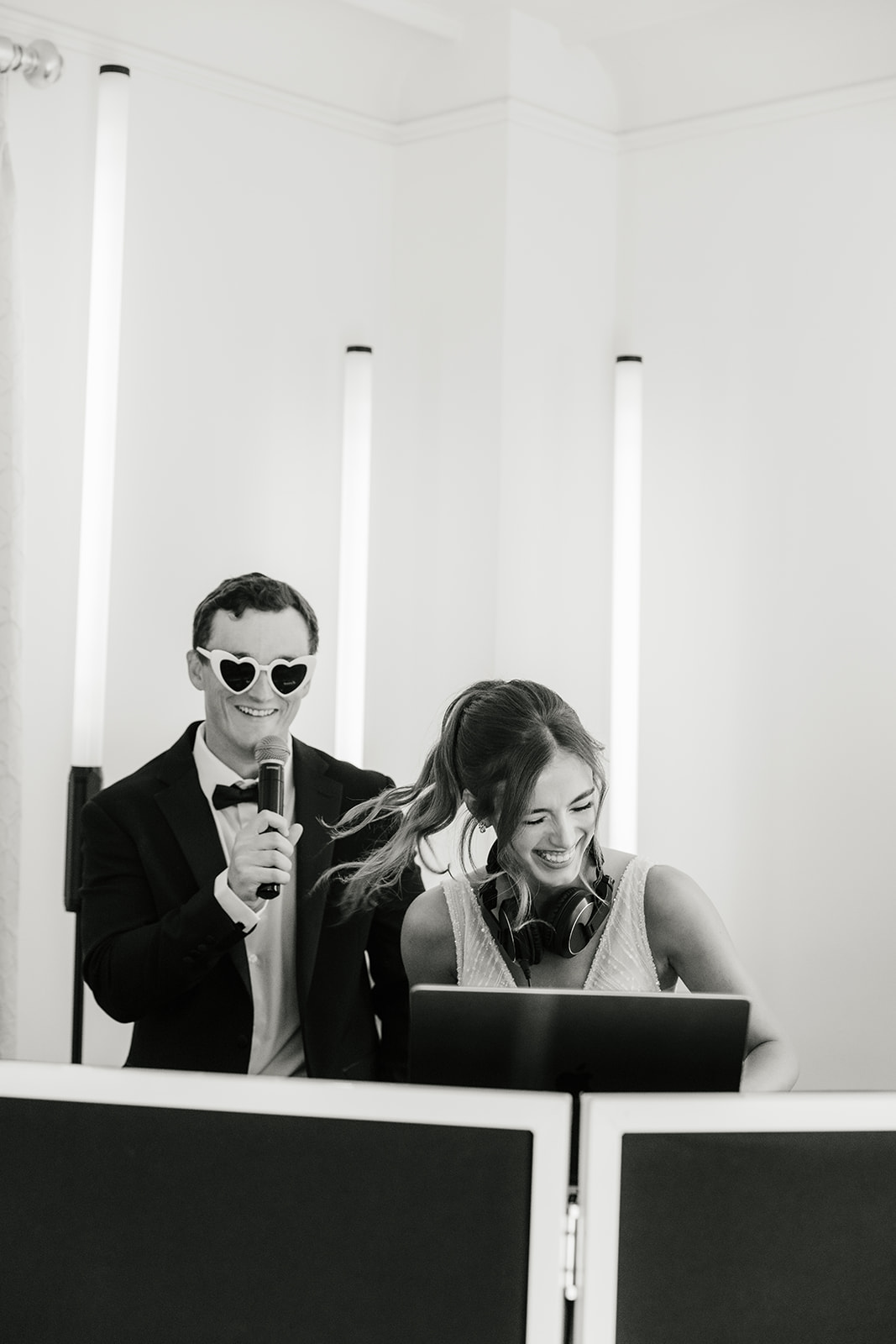 A man in a suit and sunglasses holds a microphone behind a smiling woman with headphones, both standing at DJ equipment in a brightly lit room.