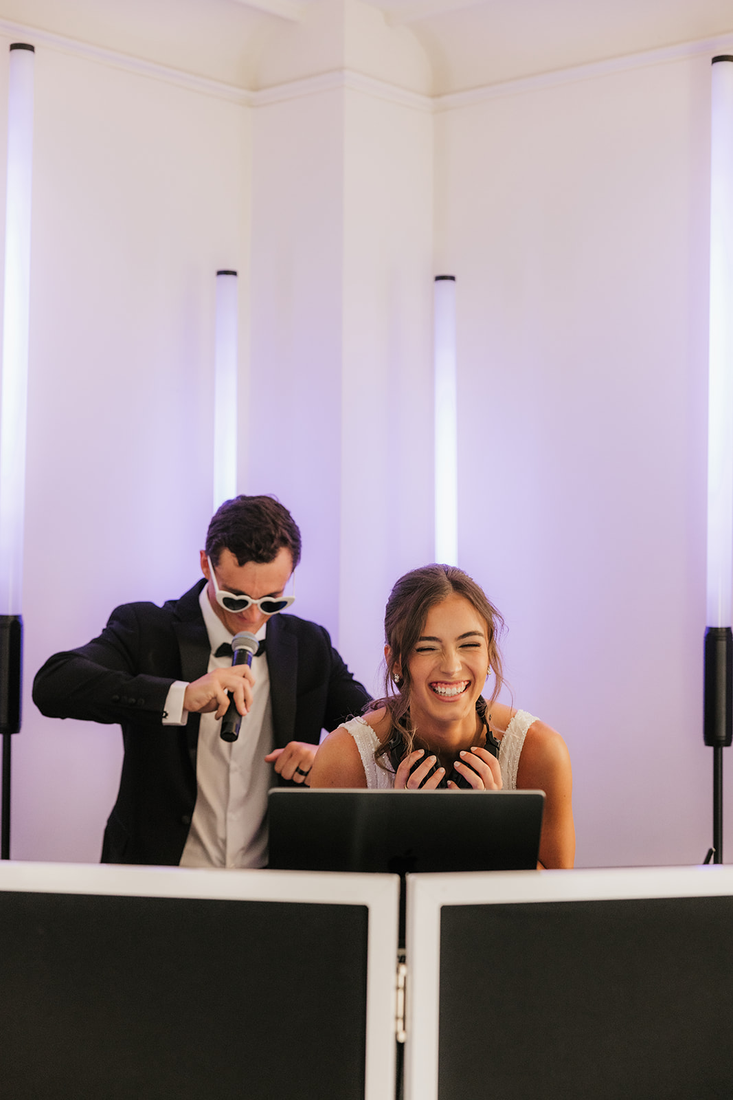 A man in a suit and sunglasses holds a microphone behind a smiling woman with headphones, both standing at DJ equipment in a brightly lit room.