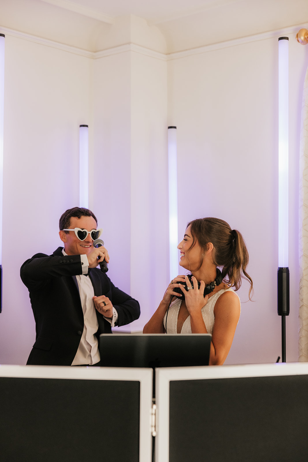 A man in a suit and sunglasses holds a microphone behind a smiling woman with headphones, both standing at DJ equipment in a brightly lit room.