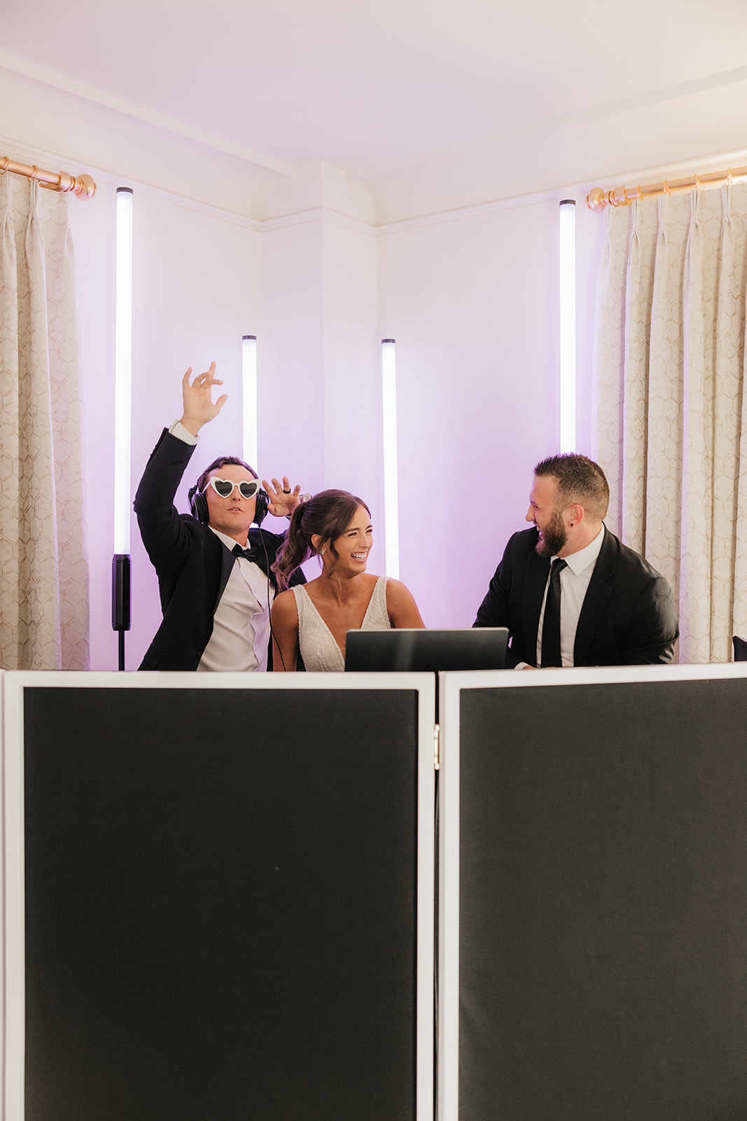 A man in a suit and sunglasses holds a microphone behind a smiling woman with headphones, both standing at DJ equipment in a brightly lit room.