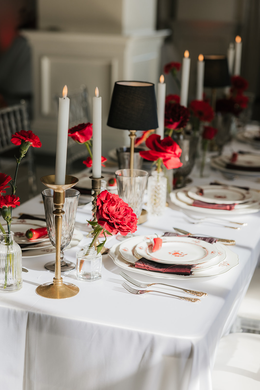 A formal table setting with stacked plates, crystal glassware, gold utensils, red napkin, a red chili pepper on the top plate, and red roses in vases on a white tablecloth for a Mayo Hotel wedding