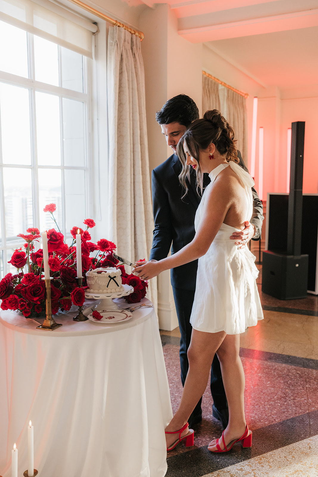 A couple stands together cutting a cake decorated with red flowers on a table with candles in a sunlit room.