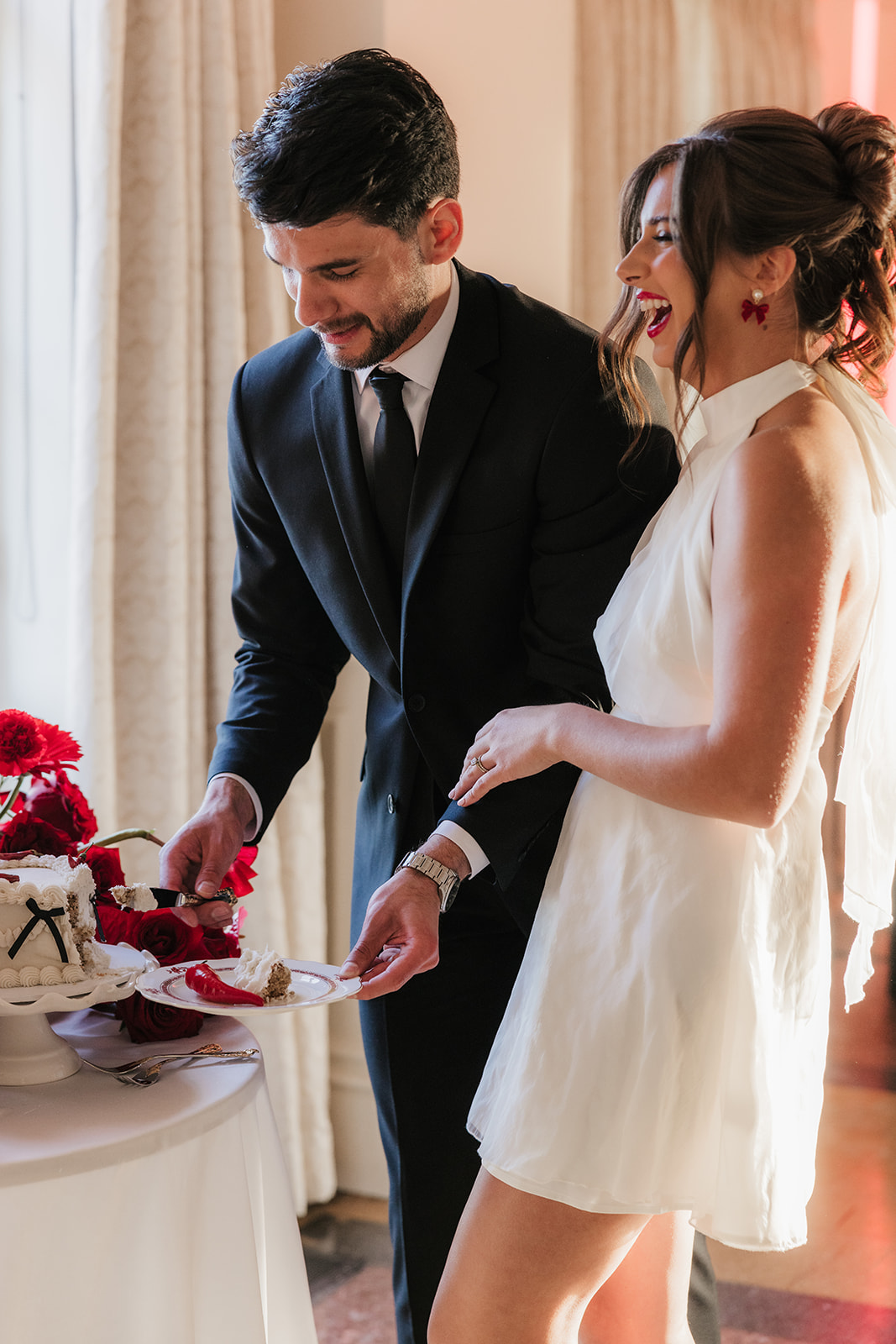 A couple stands together cutting a cake decorated with red flowers on a table with candles in a sunlit room.