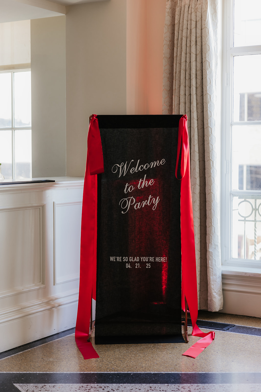 A black sign with red ribbons reads "Welcome to the Party" and is placed indoors near a window with light curtains.