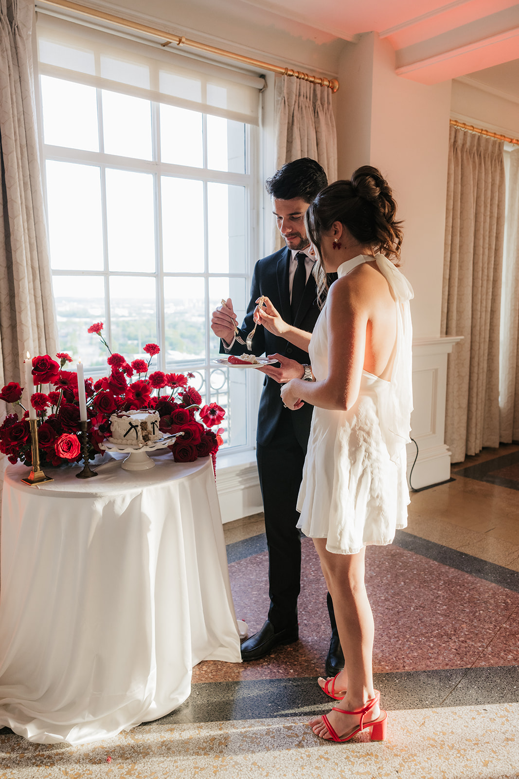 A couple stands together cutting a cake decorated with red flowers on a table with candles in a sunlit room.