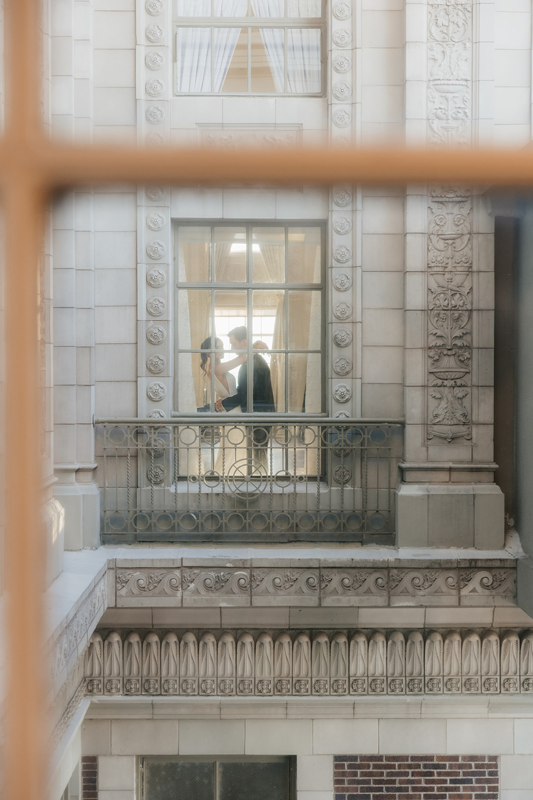 Two people stand facing each other on a balcony inside a historic building, viewed through a window from across the way.
