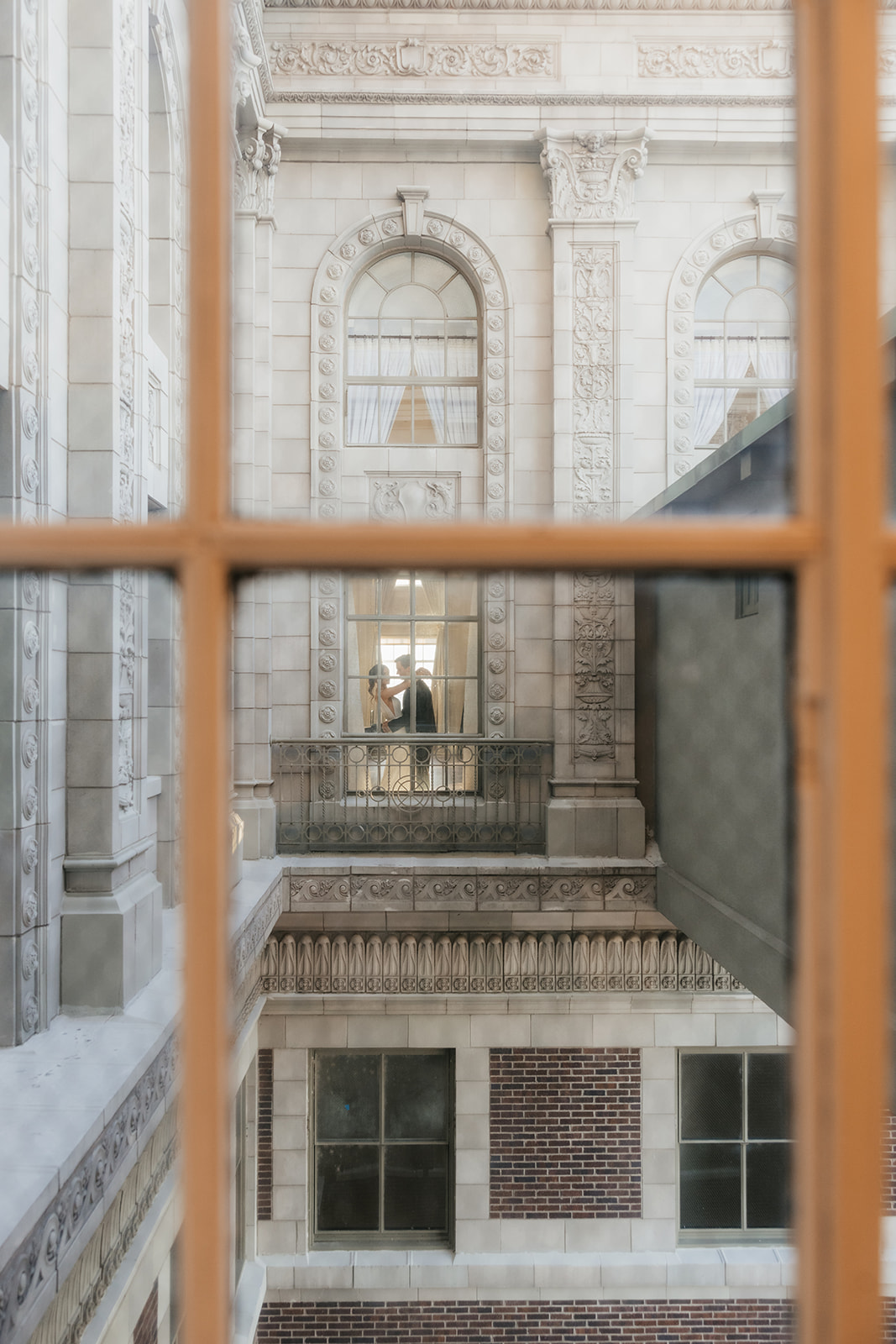 Two people stand facing each other on a balcony inside a historic building, viewed through a window from across the way.